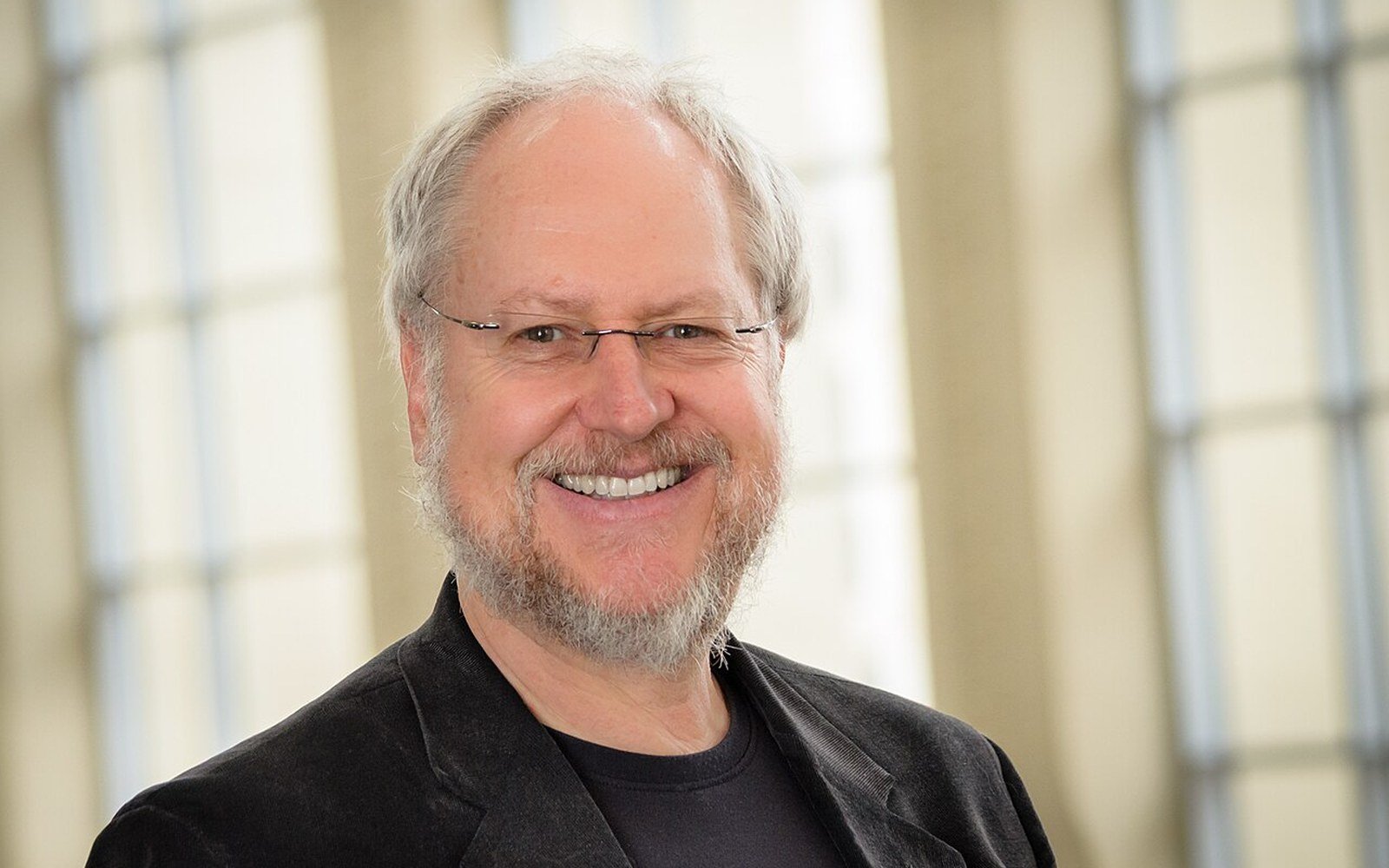 Smiling bearded man with glasses and gray hair wearing a dark blazer indoors